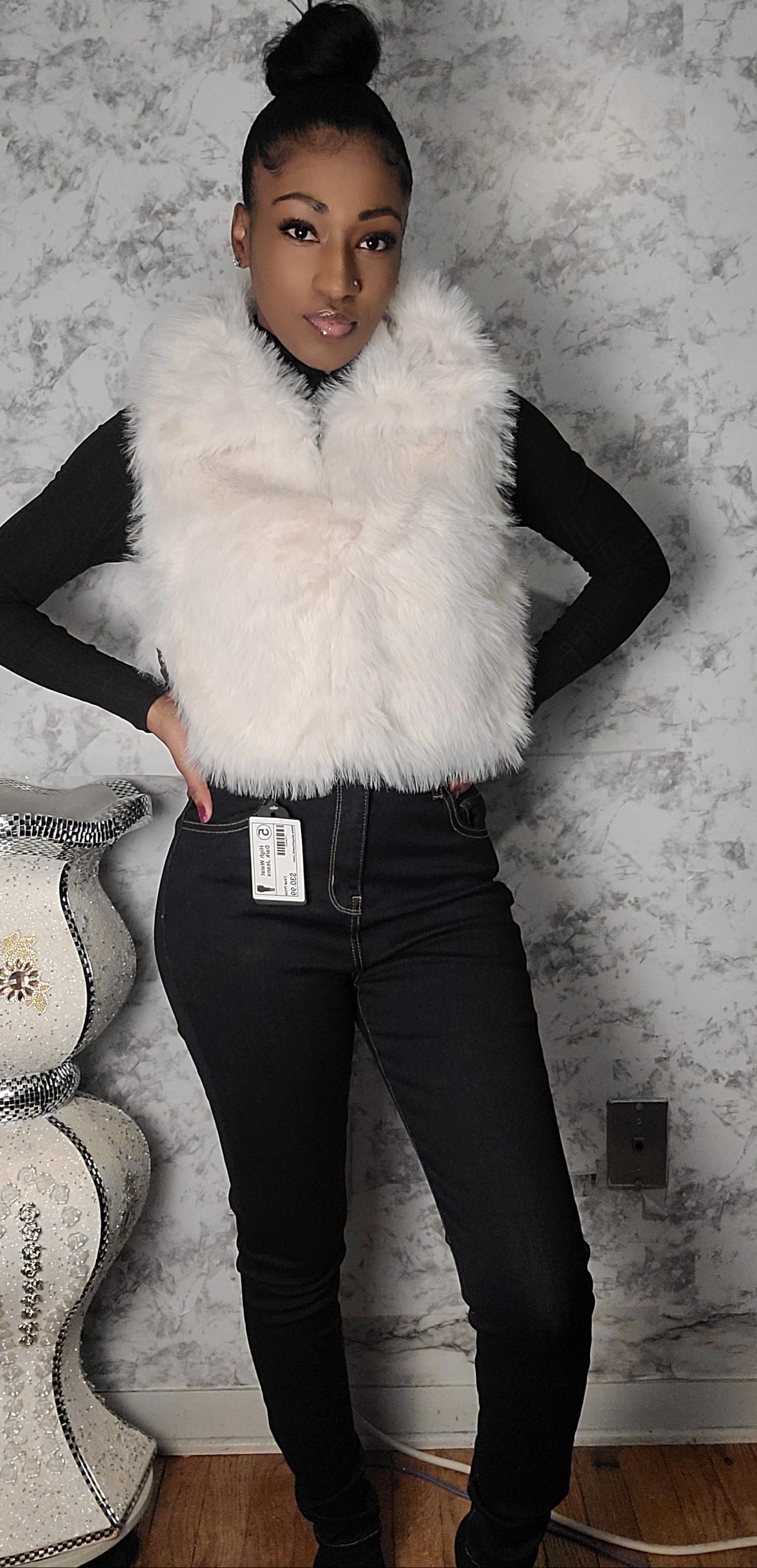 Person wearing a white fur vest over a black top and jeans, standing against a textured wall.