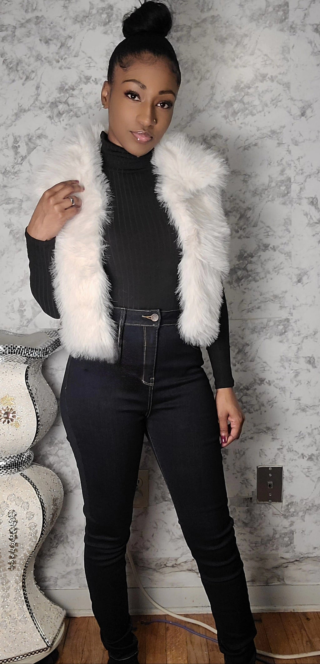 Person wearing a white fur vest over a black outfit against a textured wall.