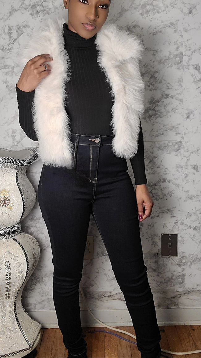 Person wearing a white fur vest over a black outfit against a textured wall. Hover Image