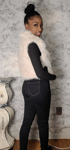 Person wearing a white fur vest over a black long-sleeve top and jeans, standing against a textured wall.