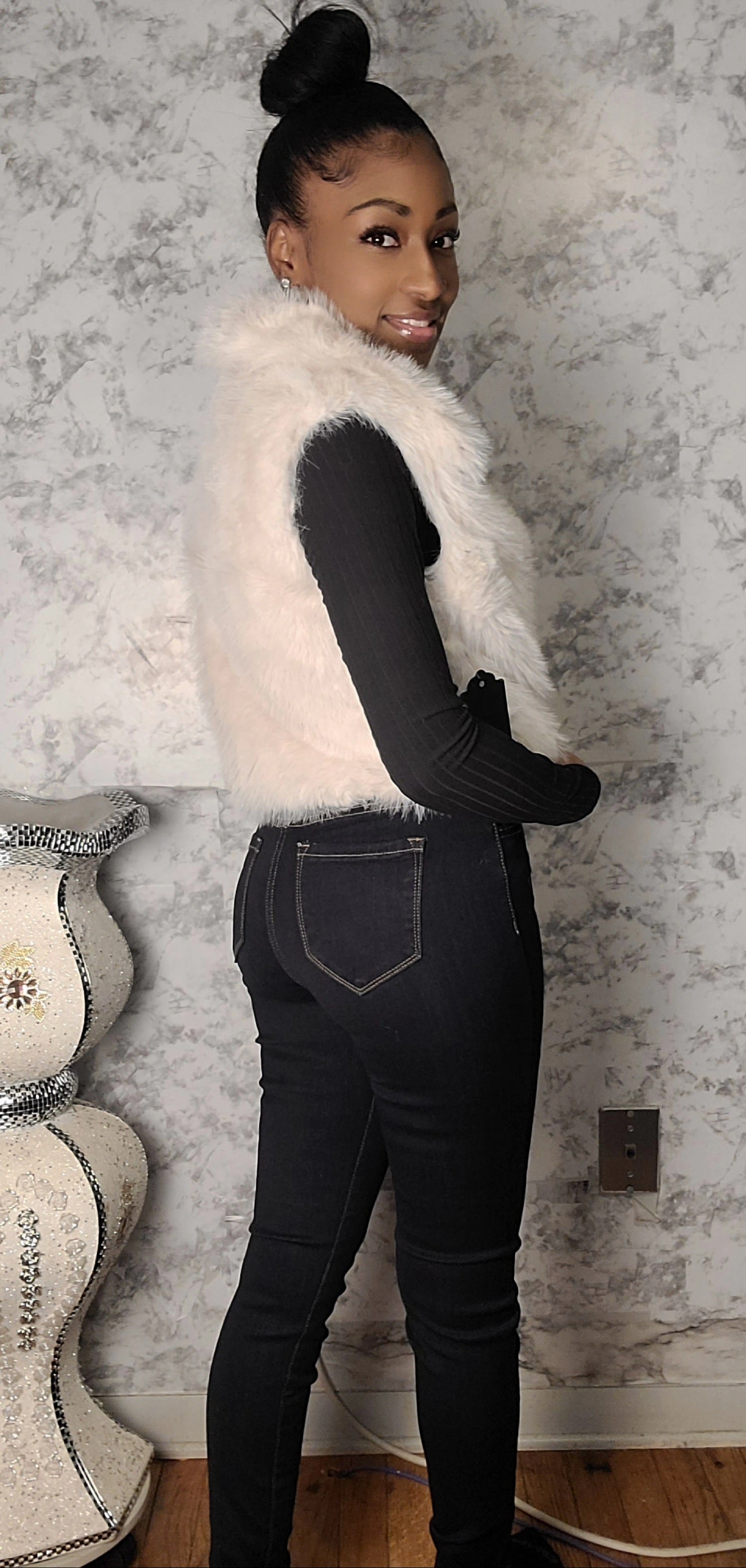 Person wearing a white fur vest over a black long-sleeve top and jeans, standing against a textured wall.