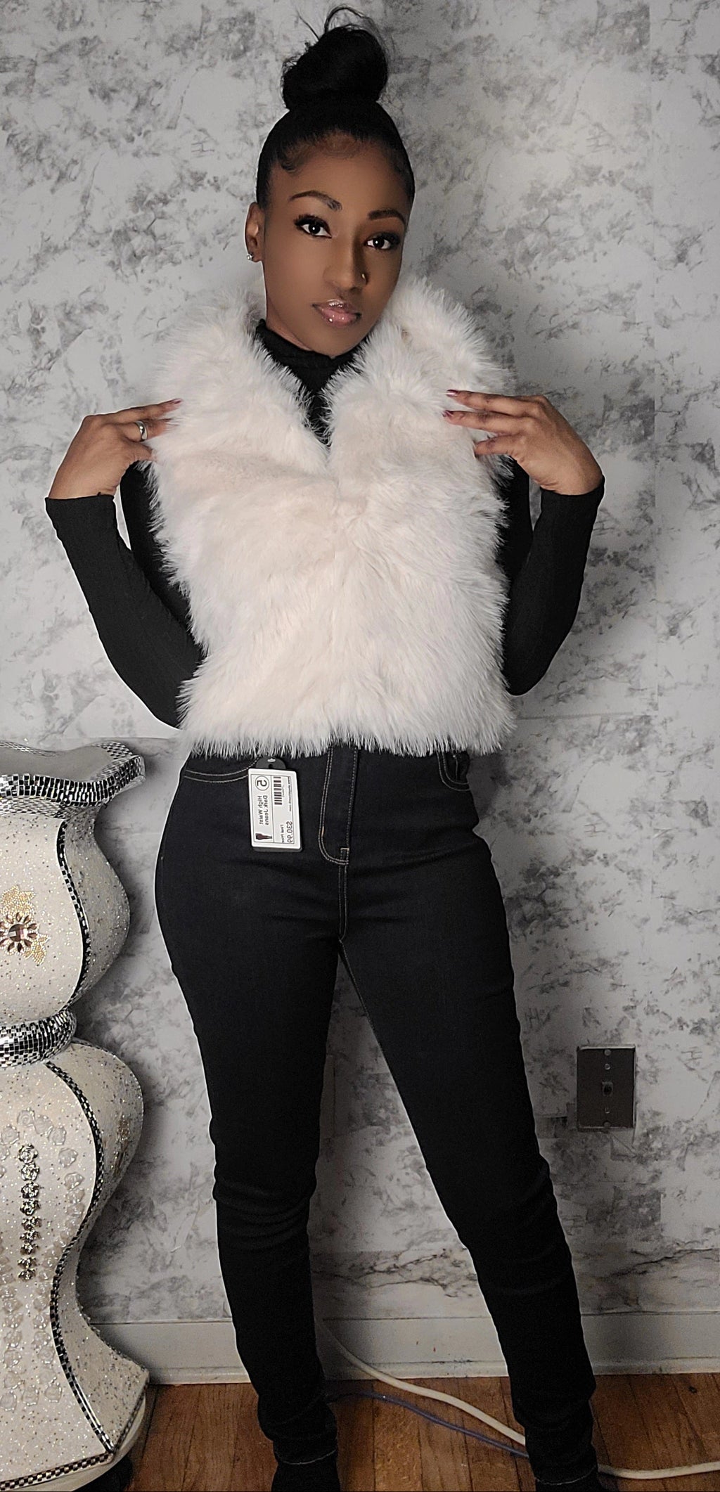 Person wearing a white fur vest over a black outfit against a textured wall.