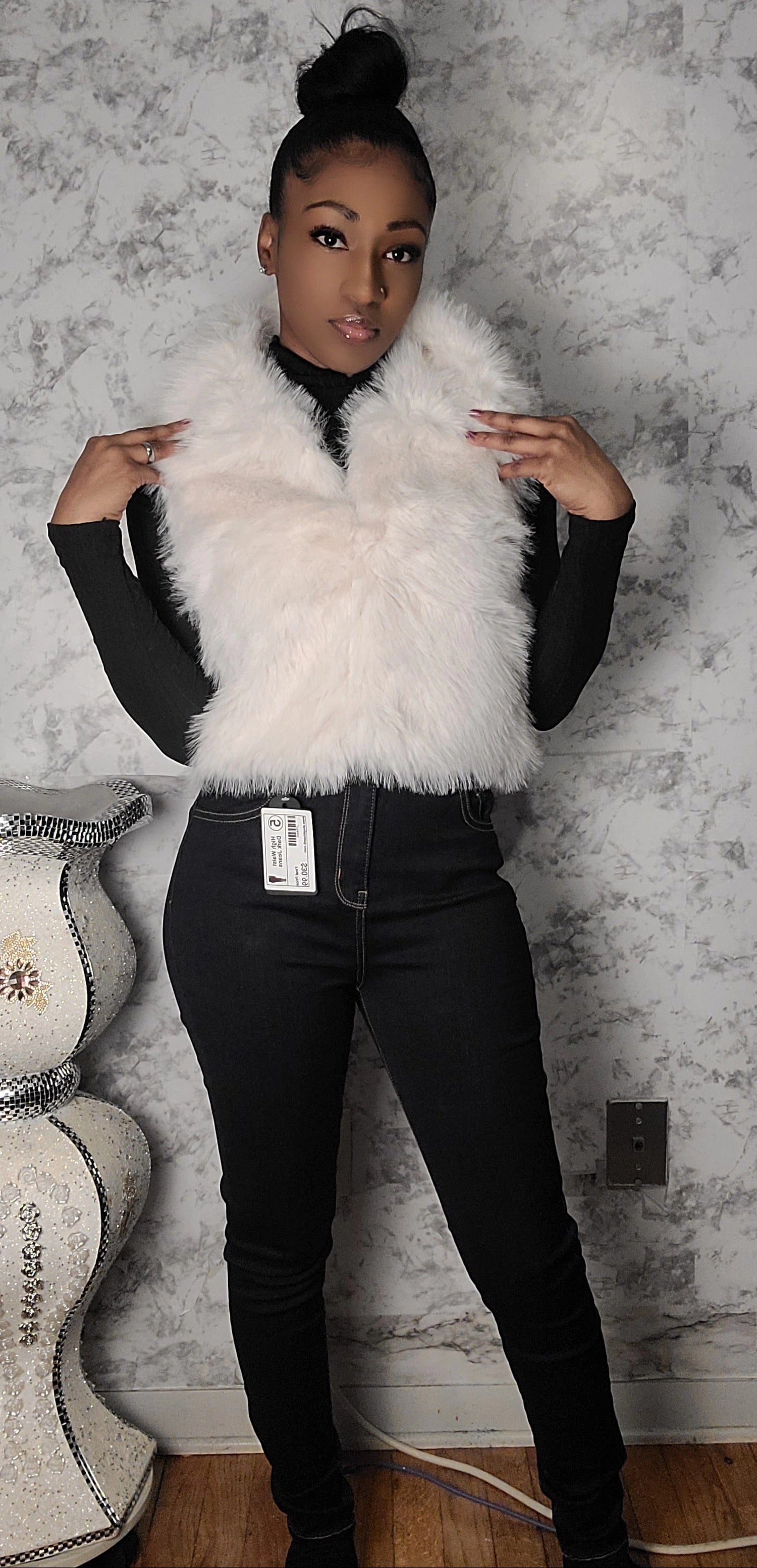 Person wearing a white fur vest over a black outfit against a textured wall.