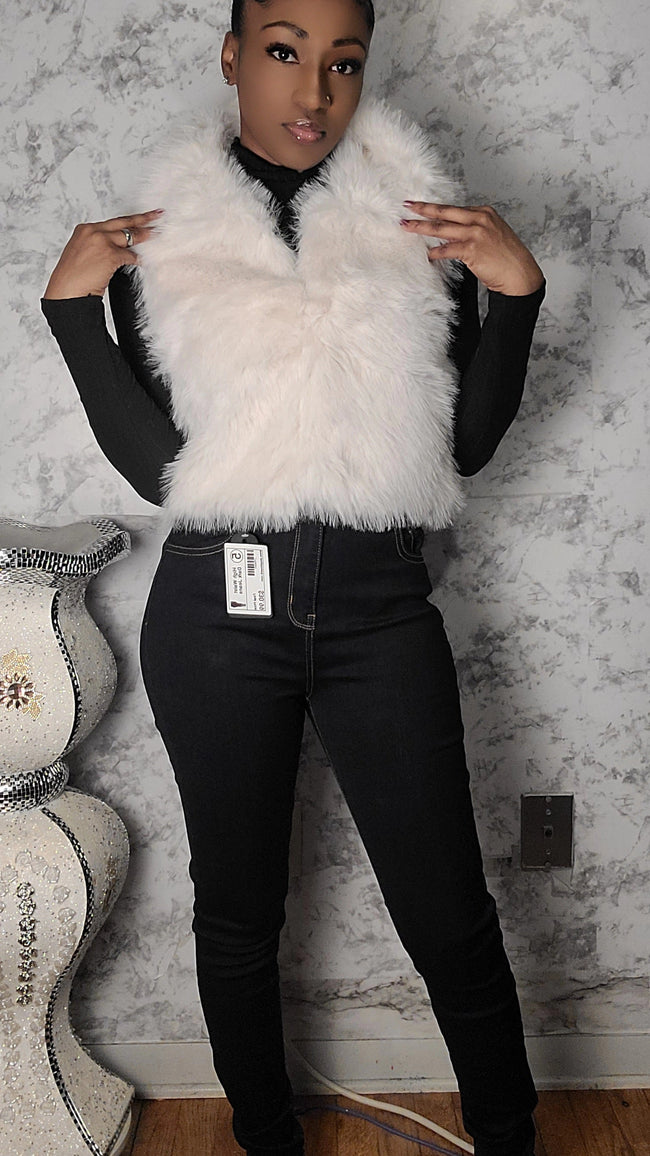 Person wearing a white fur vest over a black outfit against a textured wall. Hover Image