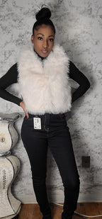 Person wearing a white fur vest over a black top and jeans, standing against a textured wall.