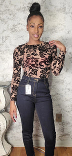 Woman wearing a sheer black floral top and dark jeans against a textured wall.
