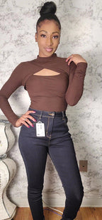 Woman wearing a brown long-sleeve top with cut-out design and dark blue jeans against a textured wall.