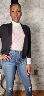 Woman wearing a black blazer, white lace top, and blue jeans against a textured wall.