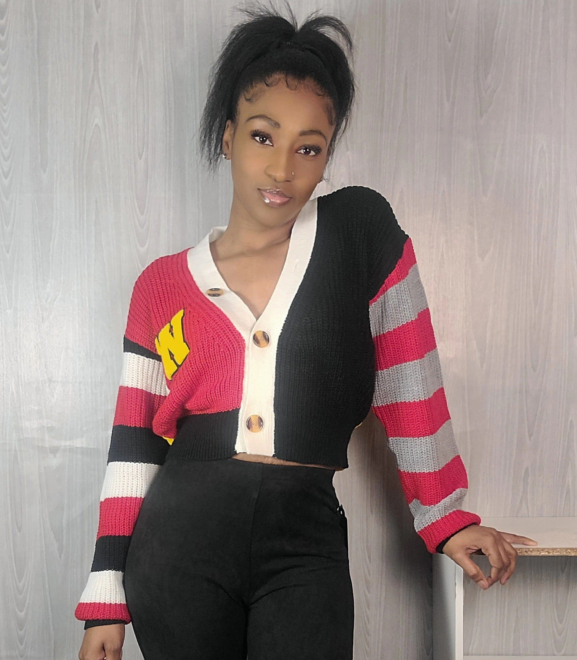 Person wearing a colorful striped cardigan with a white shirt and black pants against a light wooden panel background.