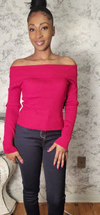 Woman wearing a bright pink off-shoulder sweater and dark jeans against a textured wall.