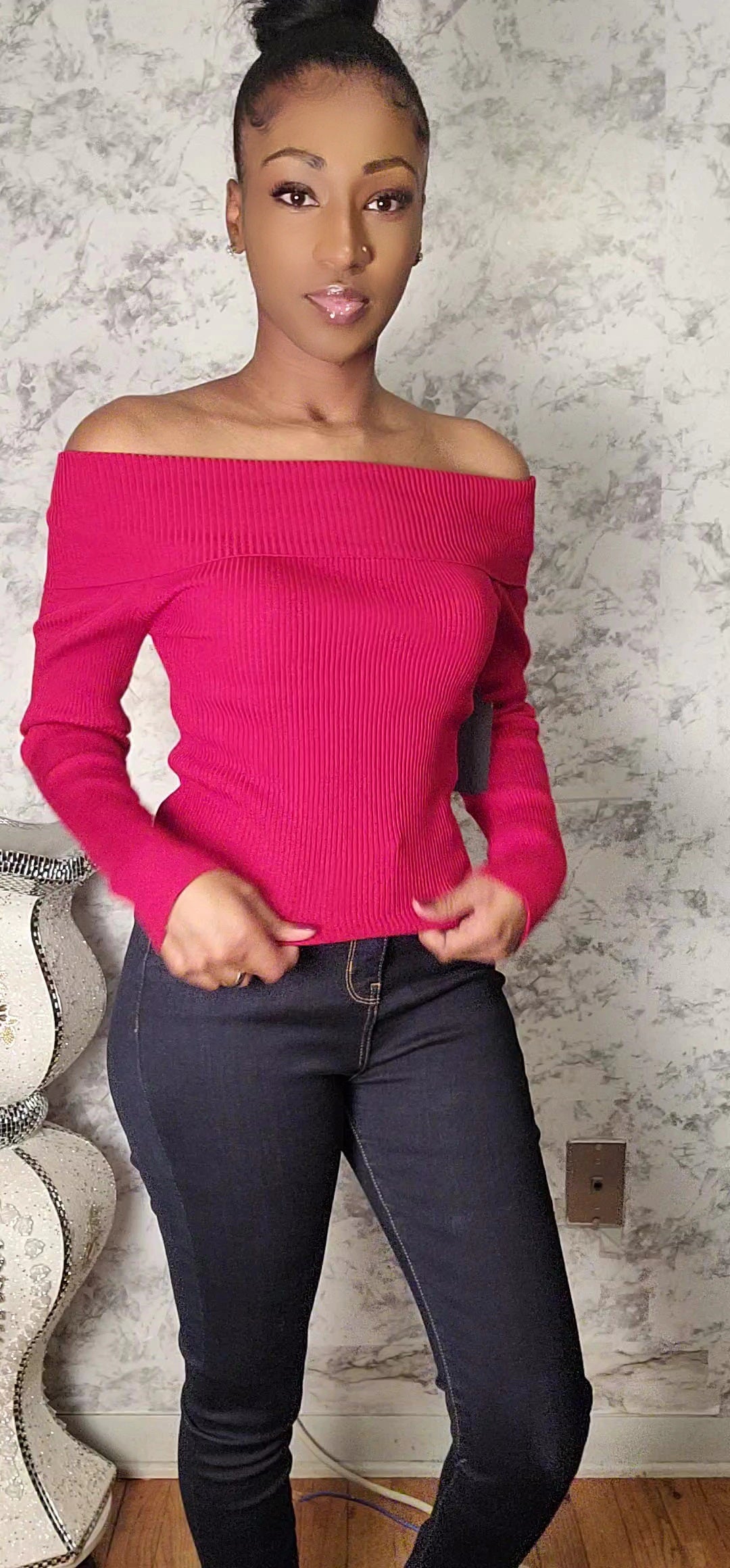 Woman wearing a bright pink off-shoulder sweater and dark jeans indoors.