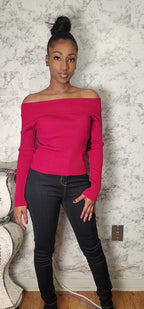 Woman wearing a bright pink off-shoulder top and black jeans against a textured wall.