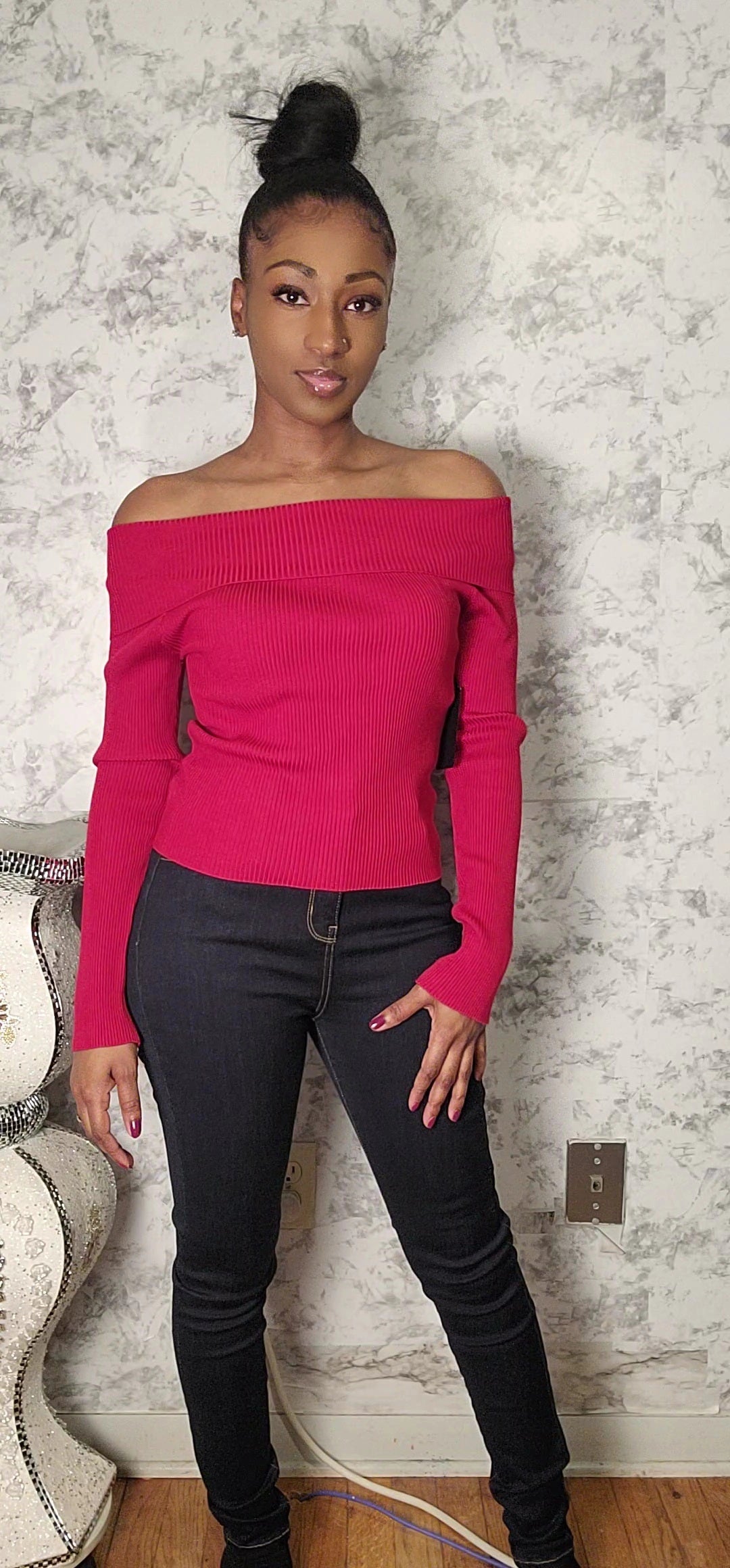 Woman wearing a bright pink off-shoulder top and black jeans against a textured wall.